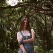 A person with long brown hair wearing a white tshirt and blue denim overalls in a woods